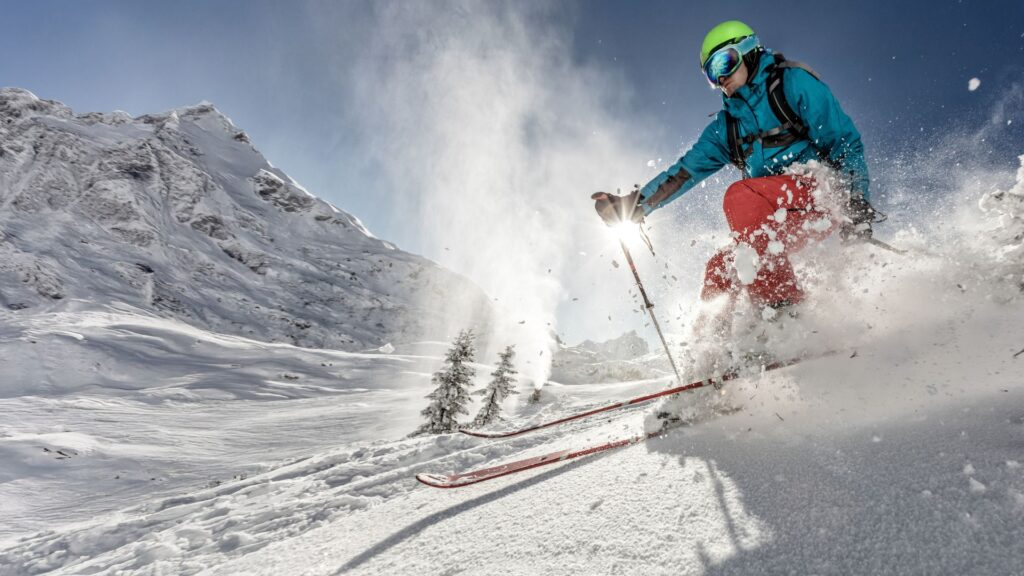 A skier in blue and red gear skiing down a snowy mountain slope, kicking up powder, with mountains and trees in the background.