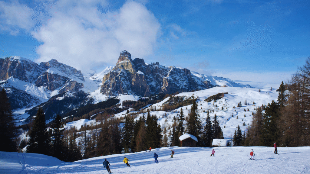 Snowy alpine landscape with skiers and snowboarders on a slope, surrounded by snow-covered mountains, evergreen trees, and a small cabin under a clear blue sky.