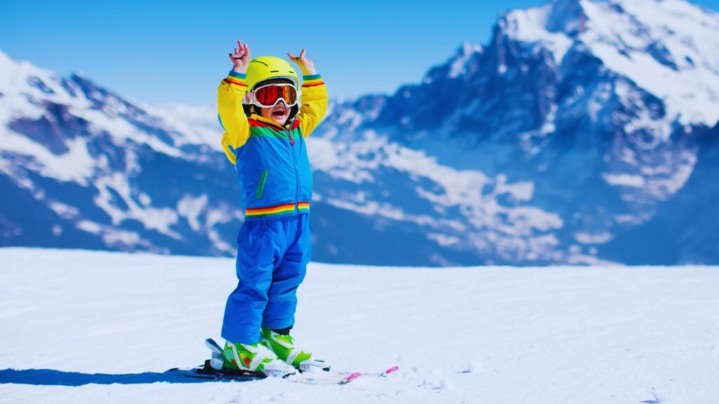 Image of a child in colorful ski gear with raised arms on a snowy slope, standing on skis against a backdrop of snow-covered mountains under a clear blue sky.