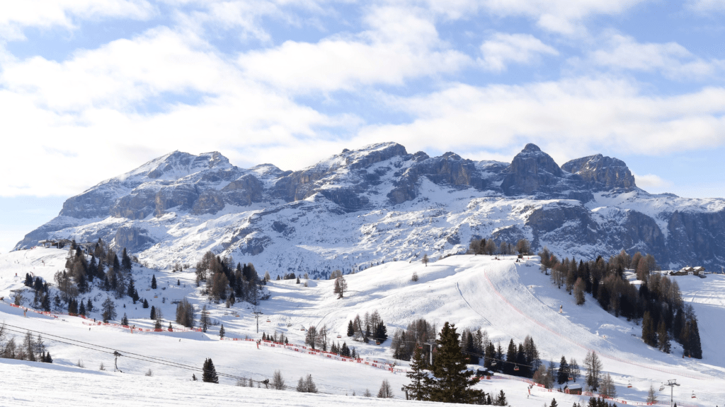 Široke zasnežene proge in dramatični vrhovi Dolomitov v Alta Badia – pogled na zasnežene gore, smučarje in gondole pod modrim nebom z rdečimi ograjami.