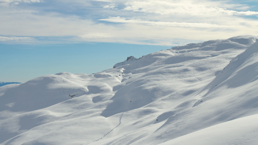 Zimski razgled na zasnežene Dolomite: mehki snežni bregovi, sledi v snegu in modro nebo z oblaki.